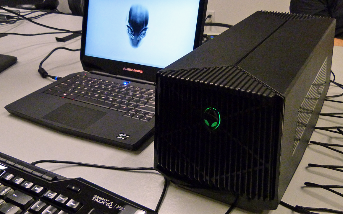 An oblong black box with a glowing alienware logo on the front next to an Alienware laptop with cords strewn on the desk.