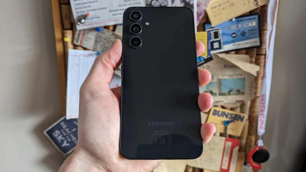 Samsung Galaxy A54 na mão em frente a um quadro de avisos