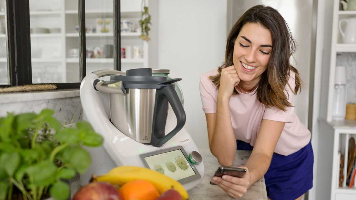 Thermomix TM6 von Vorwerk in der Küche, hübsche Frau schaut auf App und lehnt daneben, Obst und Gemüse im Vordergrund