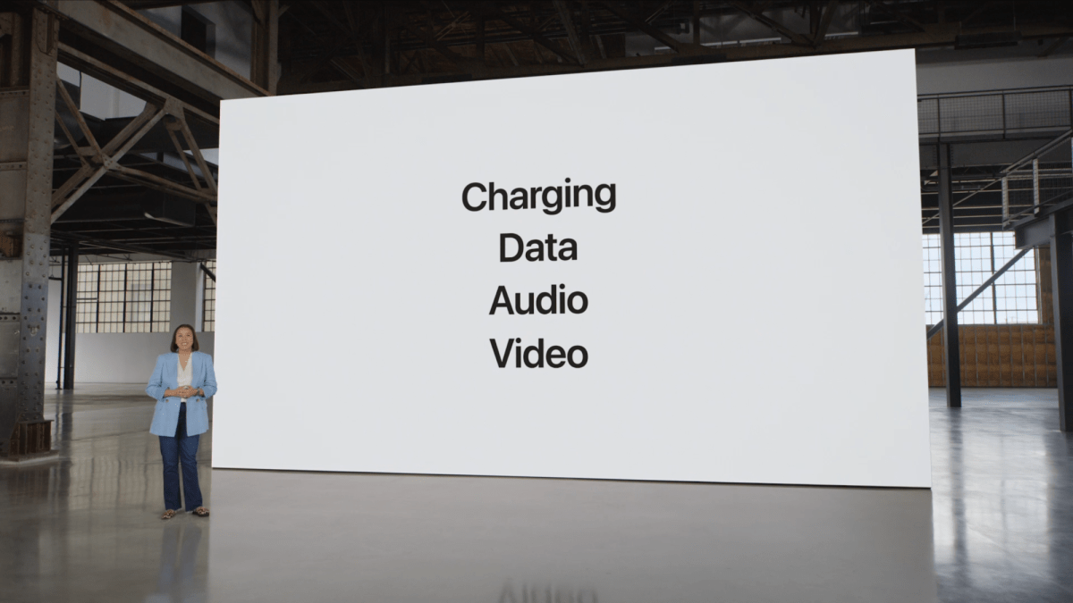 Kaiann Drance steht in einem verlassenen Lagerhaus vor einer riesigen weißen Leinwand mit den vier Worten „Charging, Data, Audio, Video“