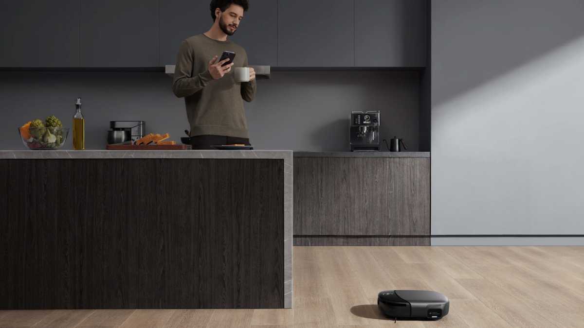 Robot vacuum cleaning a kitchen floor, with a man in the background