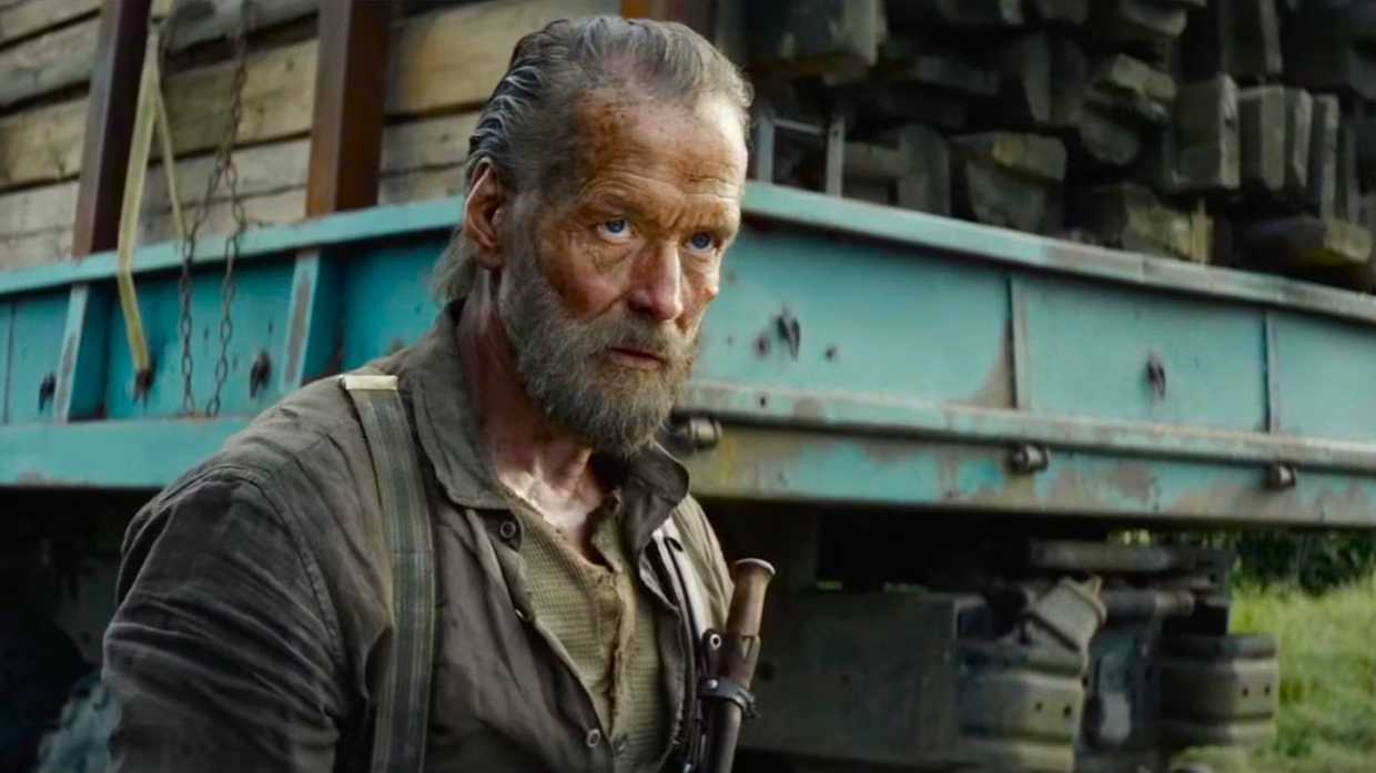 A grizzled man appears deep in thought as he overlooks a lumber-laden teal truck parked behind him.