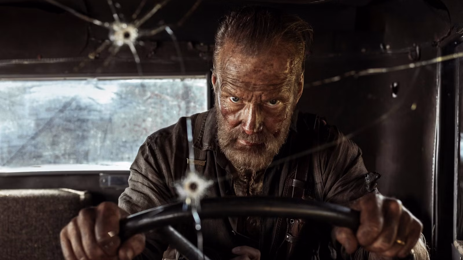 A grizzled man reflecting on the road while gazing from behind a bullet-riddled windshield