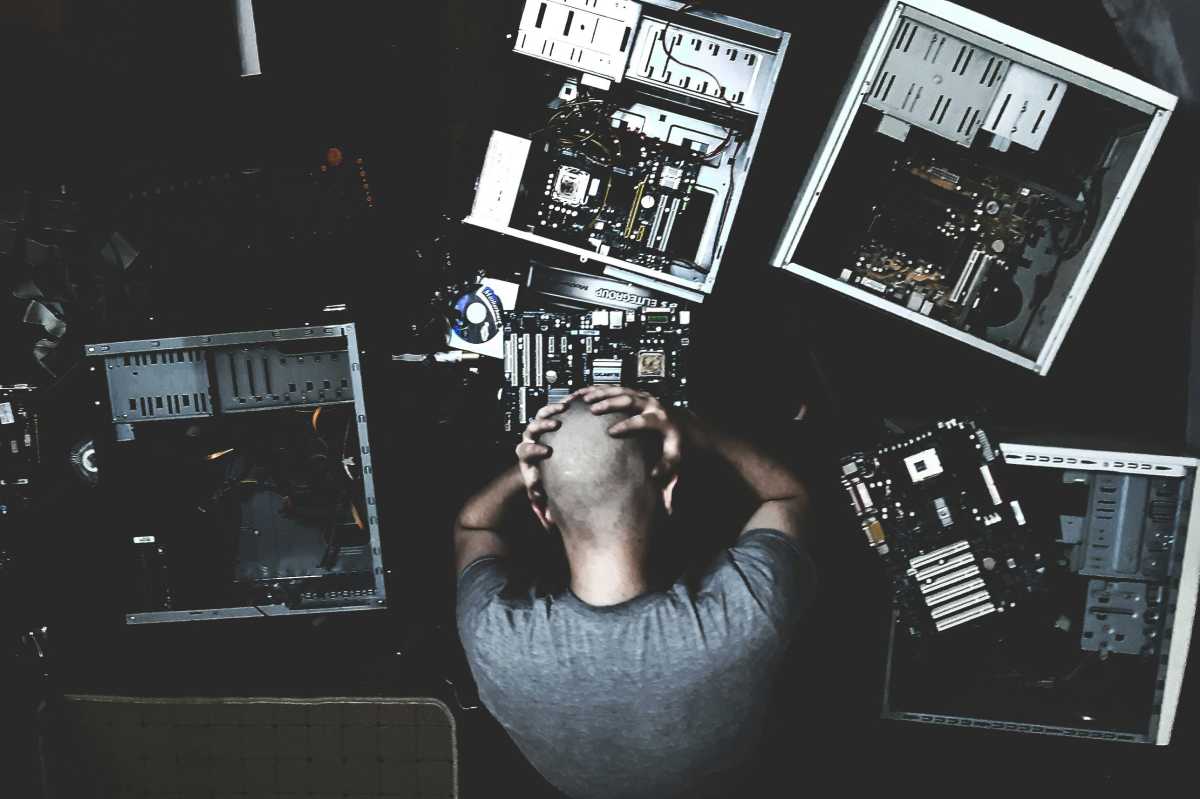 Bald person holding head in hands with multiple open PCs around them